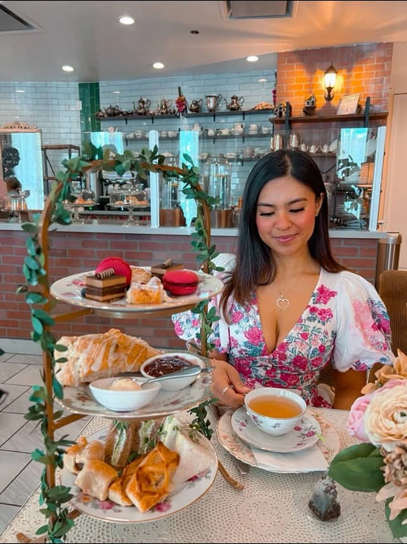 A woman in a floral dress sits at a table in a cozy cafe, enjoying afternoon tea with pastries and desserts on a three-tiered stand, holding a teacup and smiling. The background shows a decorated counter and shelves.