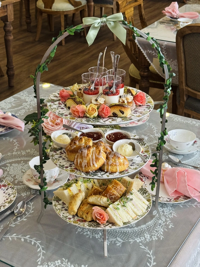 A three-tiered tray with sandwiches, pastries, scones, and desserts, decorated with roses and greenery, set on a floral tablecloth with teacups and pink napkins in a cozy tearoom setting.